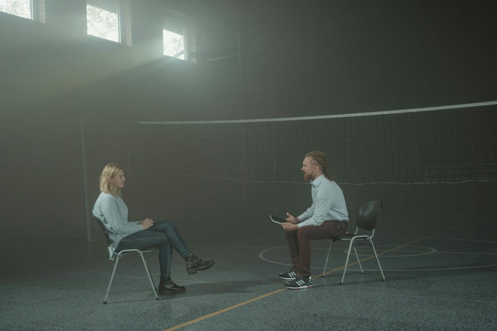 A man and a woman engage in a therapy session in a smoky indoor setting, sitting apart in a large room.
