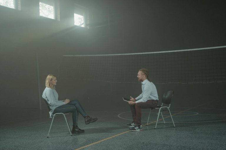A man and a woman engage in a therapy session in a smoky indoor setting, sitting apart in a large room.